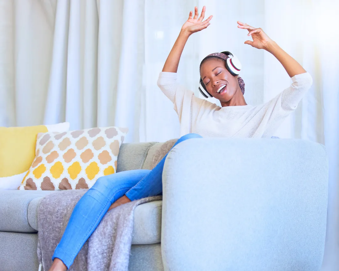 Woman relaxing on a sofa at home with headphones on, arms raised and smiling
